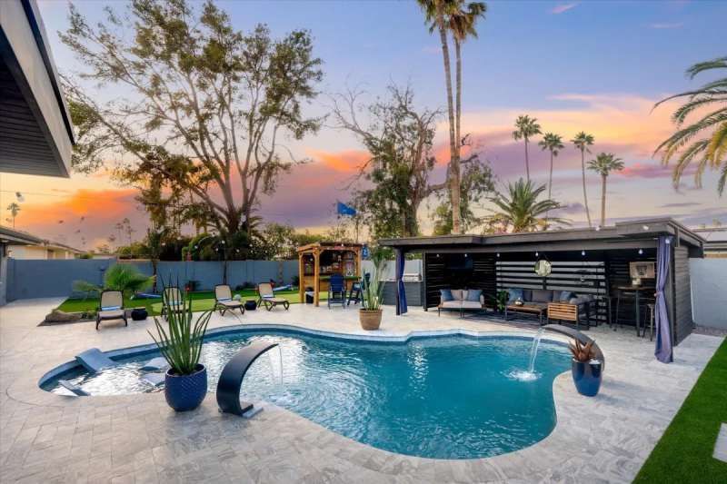 Backyard pool with a waterfall feature, patio seating, and a bar under a covered area at sunset.