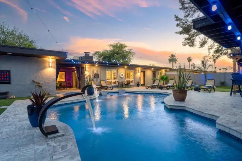 Poolside evening scene: blue pool with fountain, lounge chairs, and house with lit exterior.