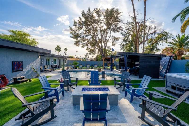 Backyard with pool, fire pit, lounge chairs, and buildings under a sunny sky.