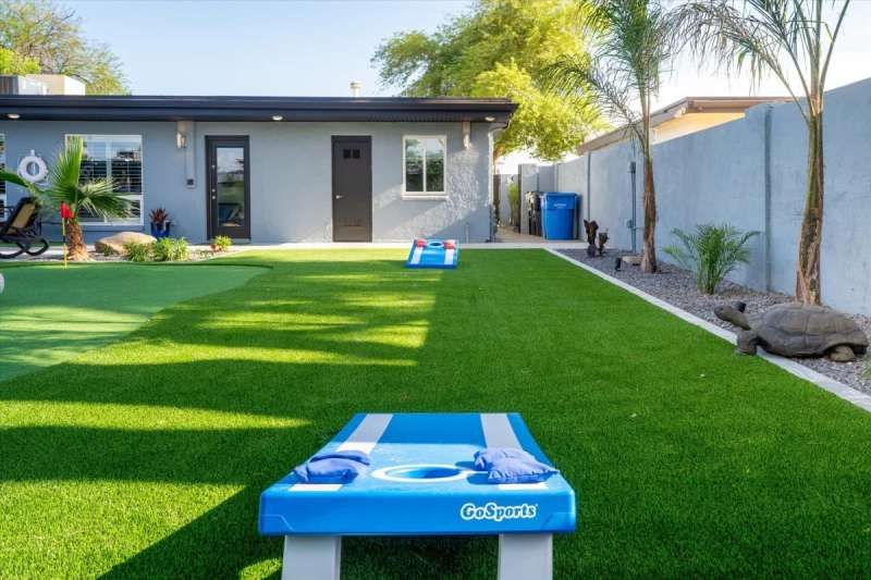 Backyard with cornhole game on artificial turf; gray house and palm trees in background.