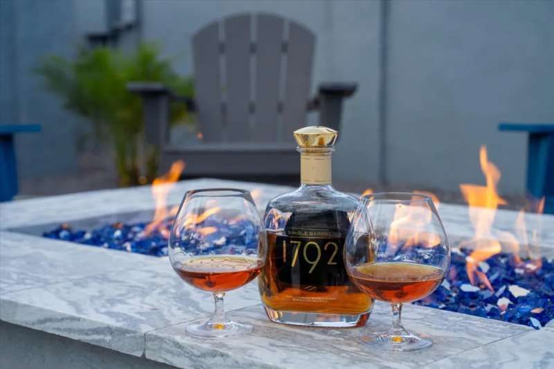 Bottle of 1792 bourbon and two glasses with amber liquid, by a fire pit with blue glass, outdoor seating in the background.
