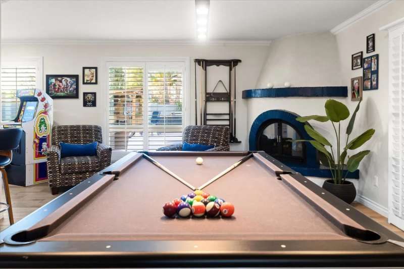 Pool table in a room with a fireplace, arcade game, and seating. Balls are racked, cue stick in place.