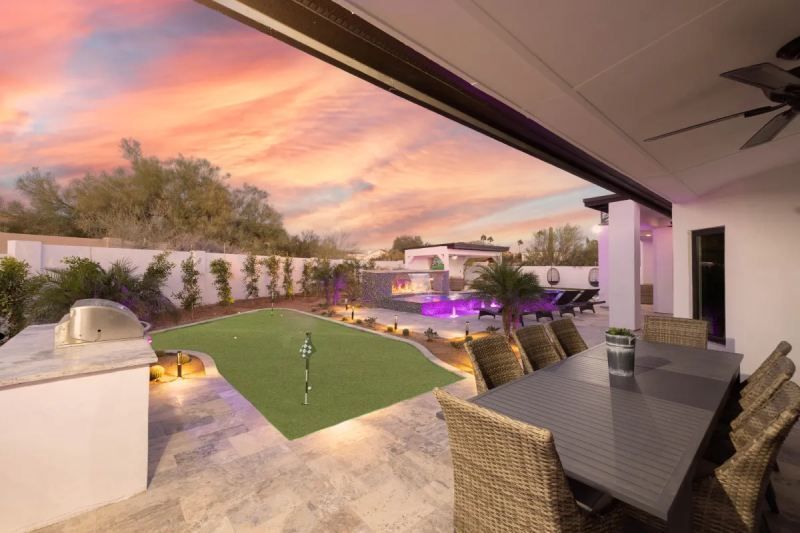 Backyard patio with dining table, putting green, pool, and sunset sky.