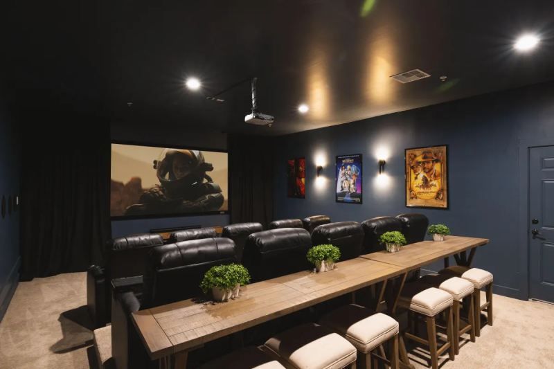 Home theater with large screen, black leather seating, and rustic table with stools. Dark walls, movie posters.