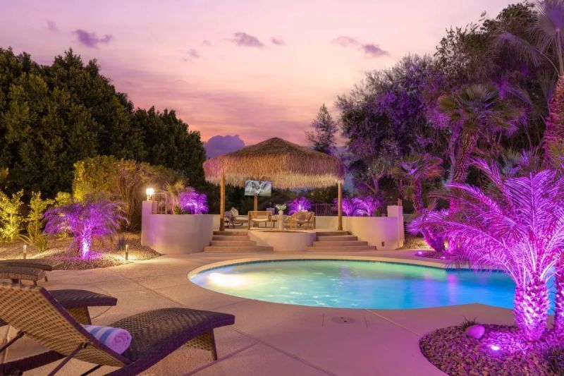 Swimming pool at dusk, illuminated by purple lights. A cabana and lounge chairs are present.