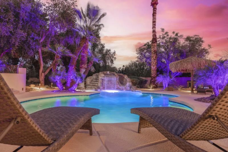 Lounge chairs face a pool with a waterfall, under purple-lit palm trees at sunset.