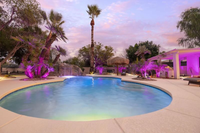 Backyard pool illuminated with pink and blue lights under a sunset sky.