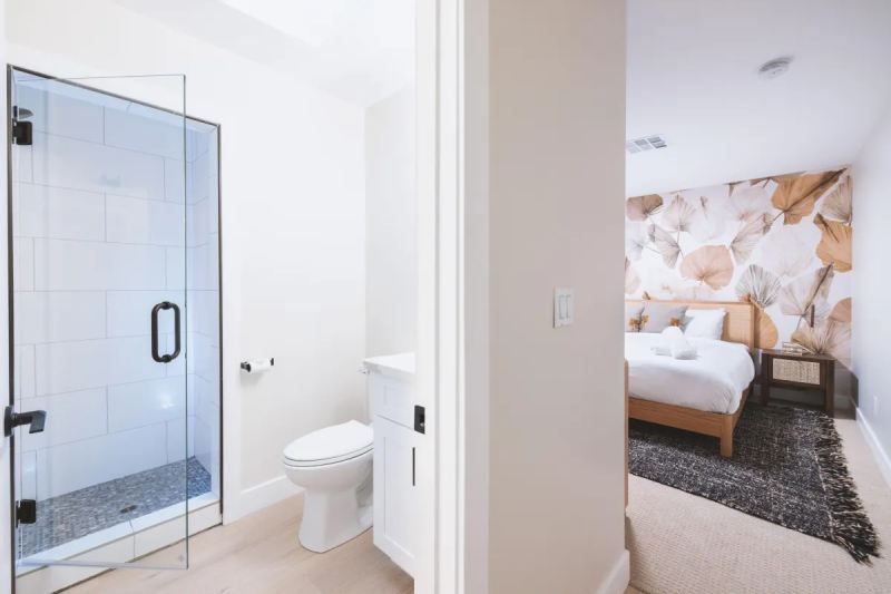 Bathroom with glass shower, toilet, and bedroom with floral wallpaper and bed.
