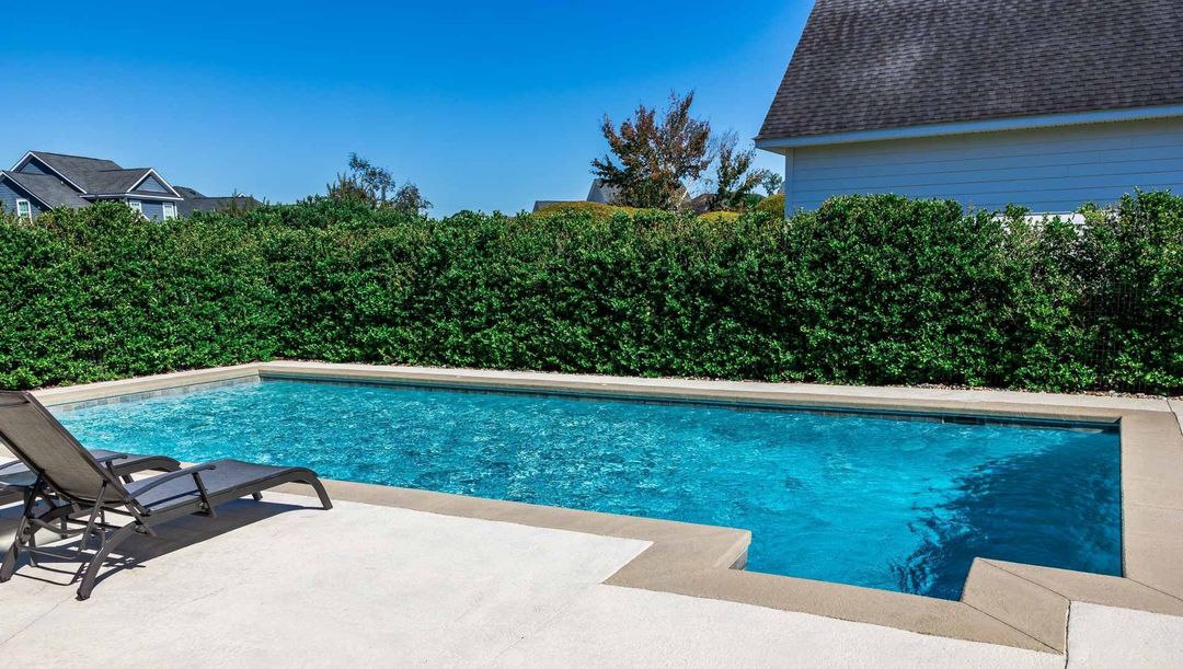 A rectangular pool with turquoise water, a lounge chair, and a hedge in a sunny backyard.