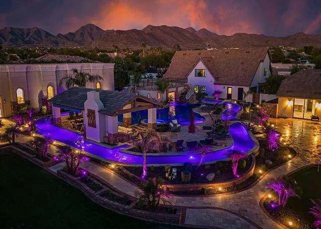 Night aerial view of a luxury estate with a curved, lit-up pool, outdoor bar, and mountain backdrop.