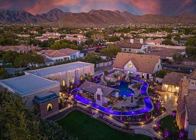 Luxury home with a winding pool lit in purple, nestled in a suburban area with mountain backdrop at sunset.