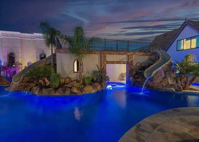 Night view of a luxurious swimming pool with slides, surrounding architecture, and blue lights.