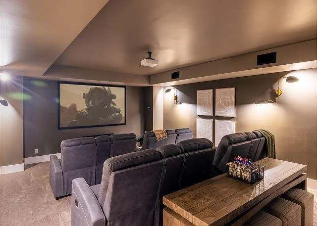 Home theater with reclining gray chairs facing a large screen, dark walls, and a wood table.