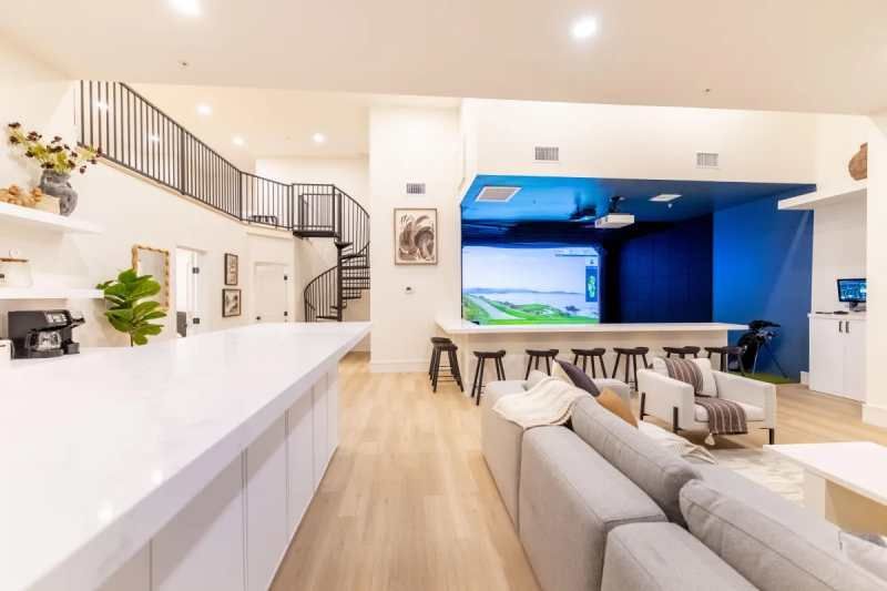 Modern interior with a golf simulator, kitchen island, spiral staircase, and gray sofa.
