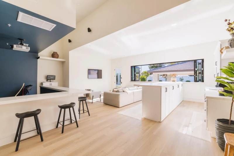 Bright, modern interior with white walls, blonde wood floors, and a blue accent wall.  A bar with stools, couch, and large windows.