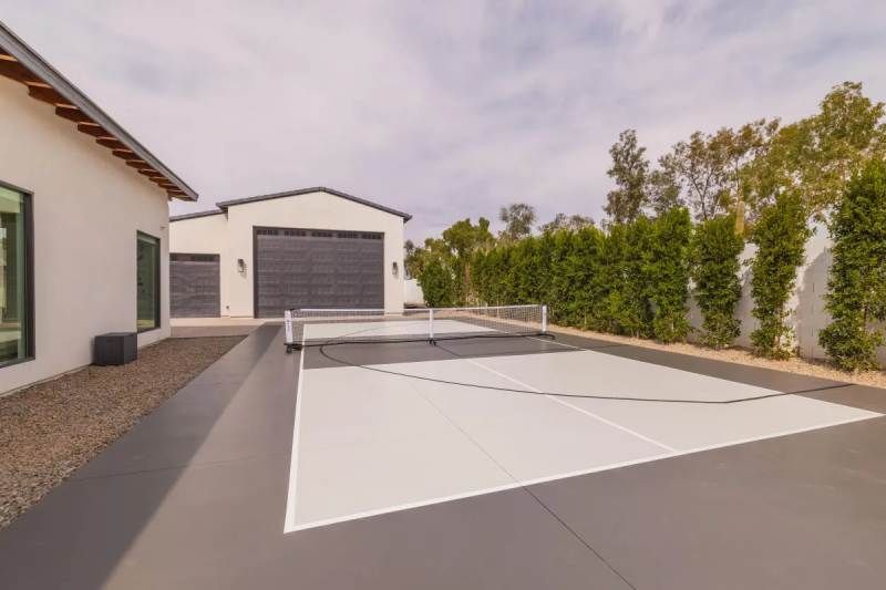 Outdoor pickleball court with black garage and white house, surrounded by trees.