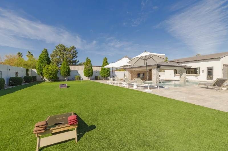 Backyard with green grass, cornhole game, patio, and pool on a sunny day.