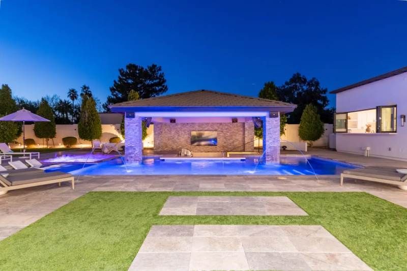 Backyard with pool, outdoor bar, and patio at dusk; blue lighting, green lawn, and stone pavers.