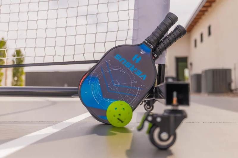 Two pickleball paddles and a ball resting against a net and a wheeled support on a court.