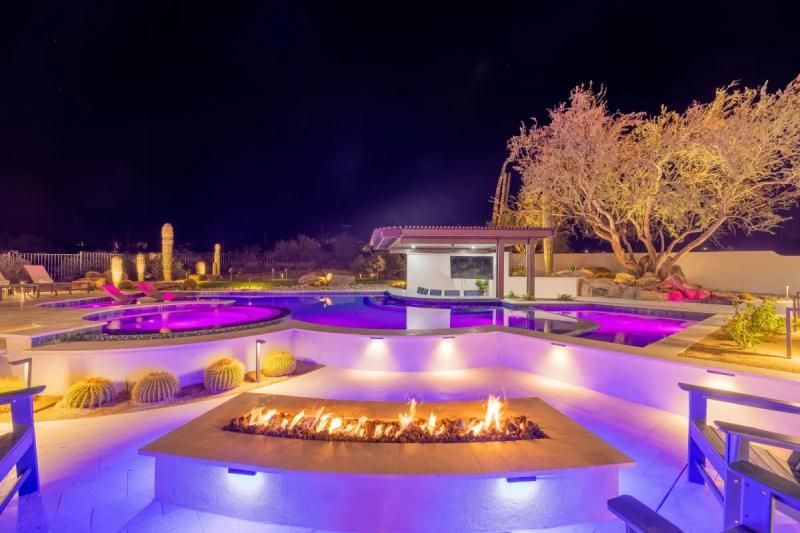 Night view of a luxurious backyard with a lit fire pit, swimming pool, and bar area.