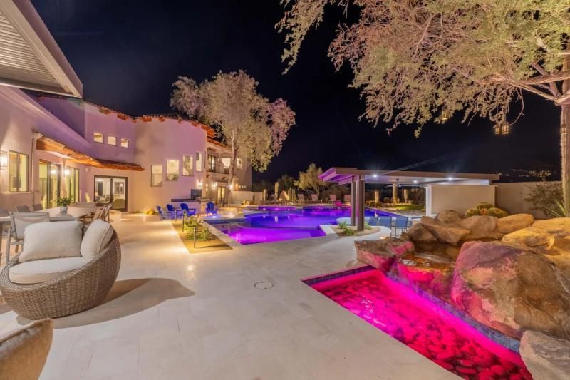Nighttime view of a luxury backyard with a pool, waterfall, and patio seating. Pink and purple pool lighting.