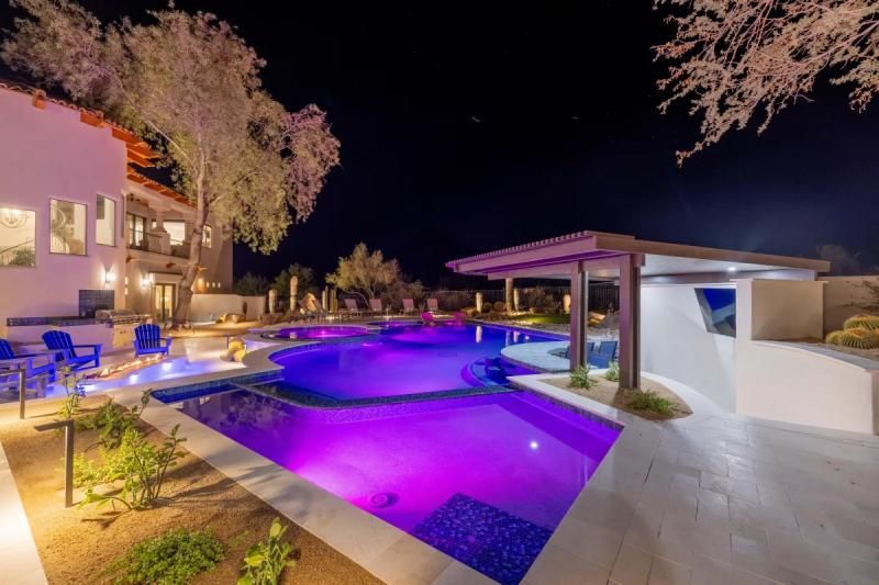 Night view of a luxurious swimming pool with purple lighting, bar area, and a large home.