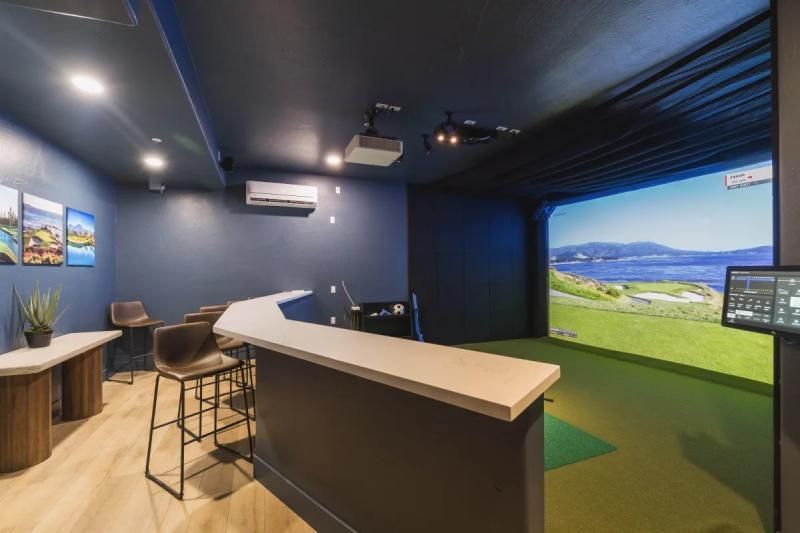 Golf simulator room with bar, screen displaying course, seating, and projector. Dark blue walls, light wood accents.