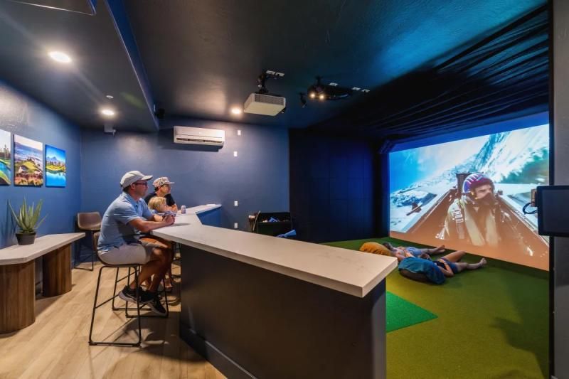 A gaming room with people watching a mountain scene projected on a screen, with a bar and benches.