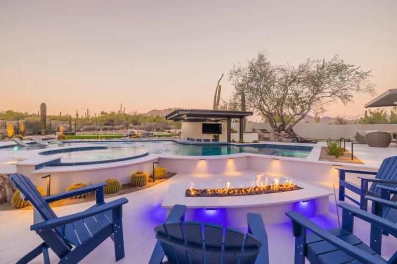 Patio with blue chairs, fire pit, pool, and outdoor bar at sunset.