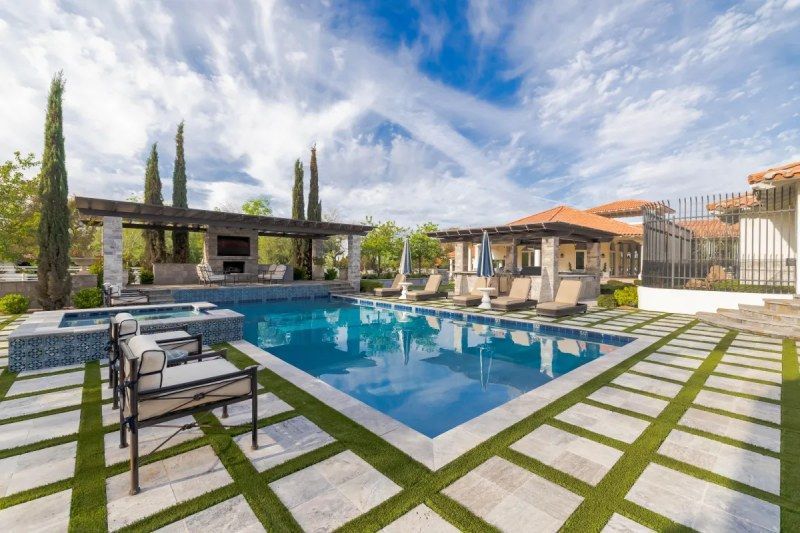 Swimming pool surrounded by stone squares and artificial turf, cabanas, and a pergola under a blue sky.