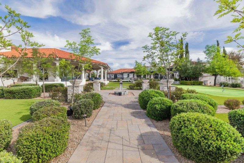 Stone walkway leading to a building with a red-tiled roof, lined with green bushes, under a blue sky with clouds.