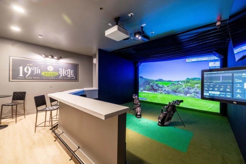 Indoor golf simulator with a bar; screen shows a golf course, clubs and monitor visible.