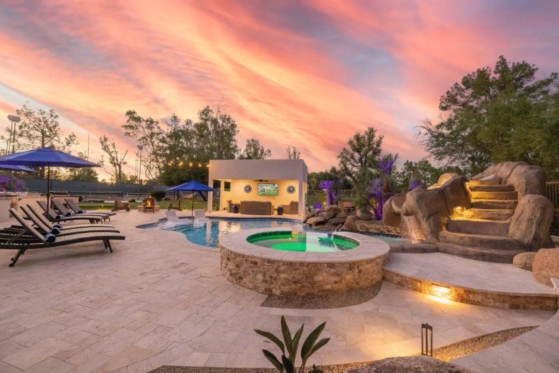 Poolside at sunset: pool, spa, lounge chairs, small bar with TV, and steps up to a rock-lined structure.