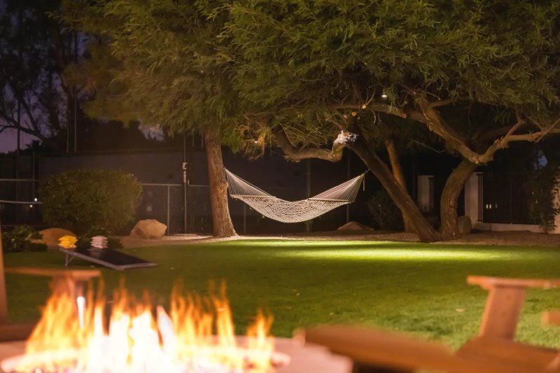 Backyard at dusk with a hammock strung between trees, illuminated lawn, and a fire pit.
