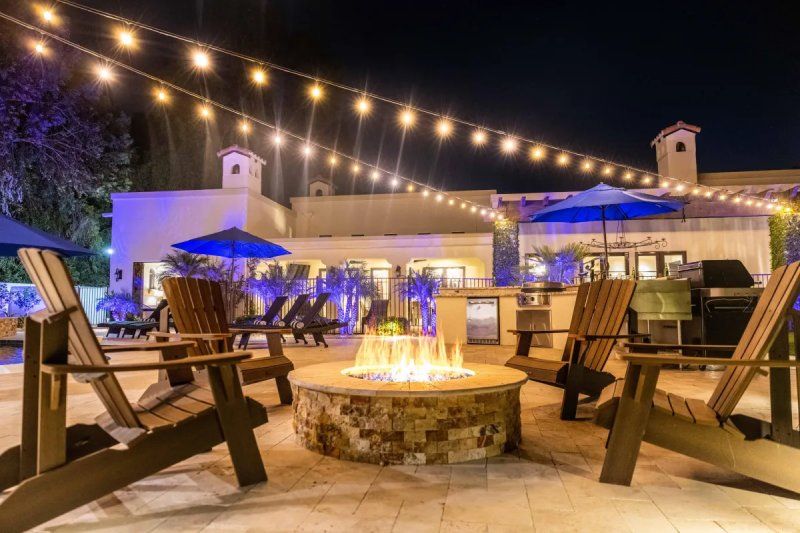 Nighttime outdoor seating area with fire pit, wooden chairs, string lights, and a building in the background.