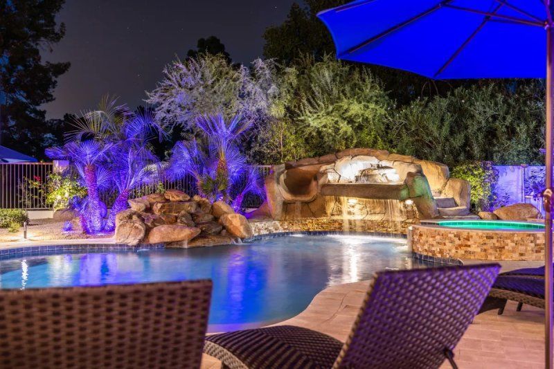 Pool with waterfall and lounge chairs at night. Blue and white lighting illuminates the scene.