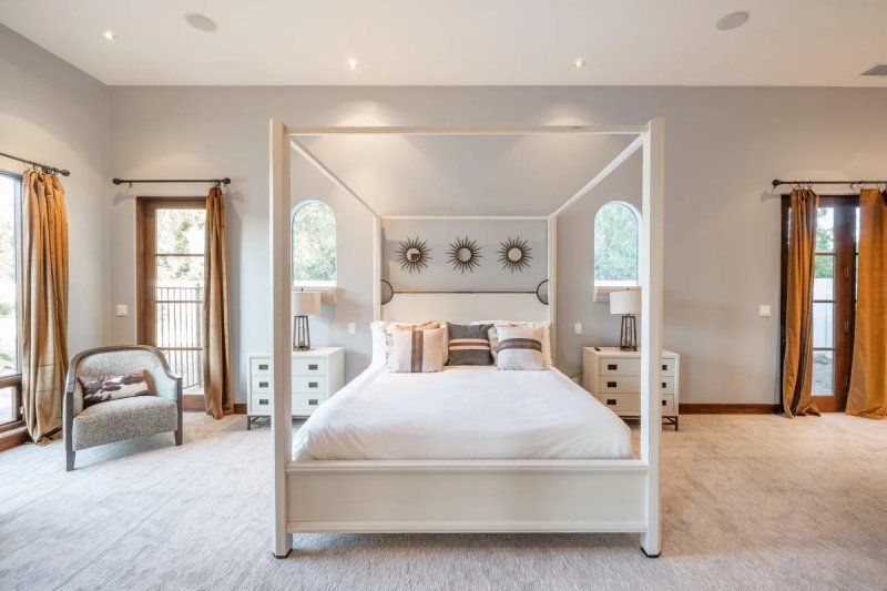 Spacious bedroom with white four-poster bed, neutral tones, and two windows with sheer curtains.