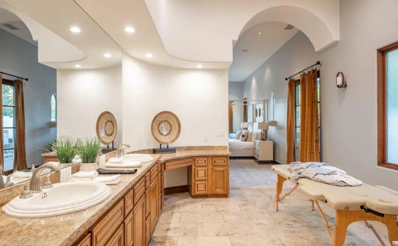 Bathroom with vanity, massage table, and open doorway to bedroom. Light-colored walls, beige tile, wood accents.
