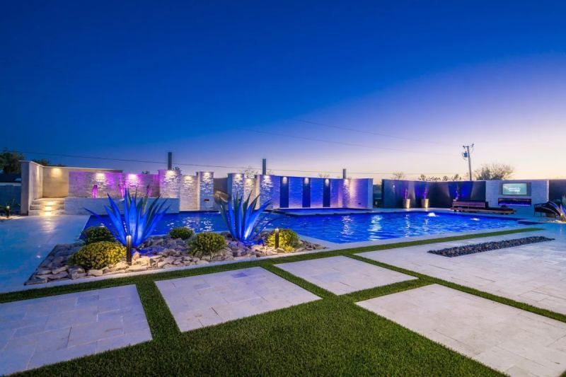 Nighttime view of a backyard pool with blue lights, a tiled waterfall wall, and large square pavers on a grassy lawn.