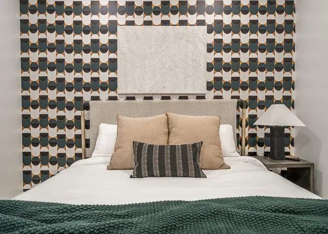 Bedroom with patterned wallpaper, bed with pillows, and a nightstand with a lamp.
