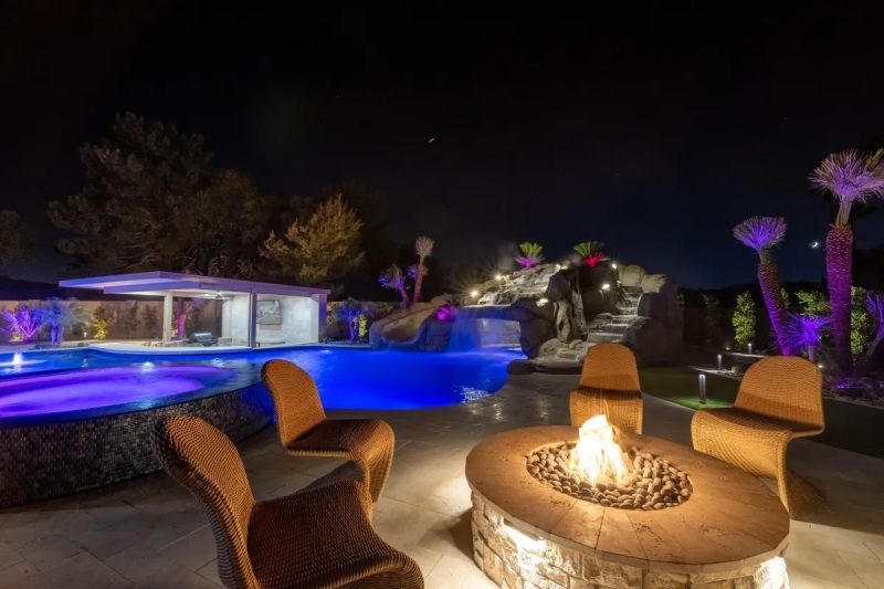 Nighttime backyard with a pool, bar, fire pit, and seating. Purple and blue lighting.