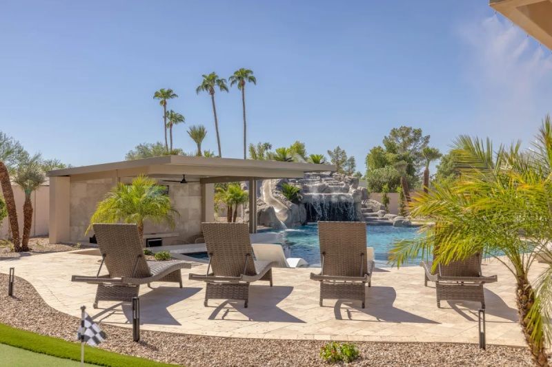 Poolside scene with lounge chairs, a waterfall, and palm trees under a blue sky.
