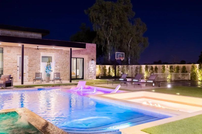 Nighttime view of a luxurious backyard with pool, spa, and house illuminated with blue and orange lights.