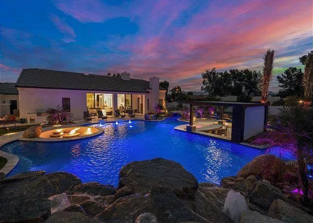 Luxury home pool at sunset, illuminated blue, with fire pit, swim-up bar, and colorful sky.