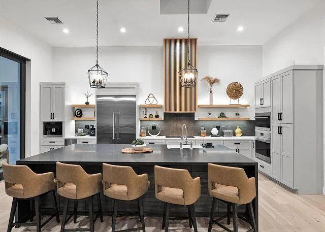 Modern kitchen with dark island and tan bar stools. Stainless steel appliances, gray cabinets, and wooden accents.
