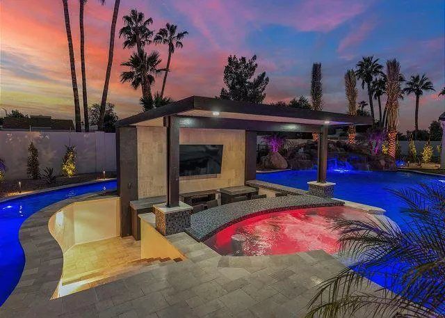 Luxury outdoor pool with swim-up bar, lights, seating, and a TV, at dusk.