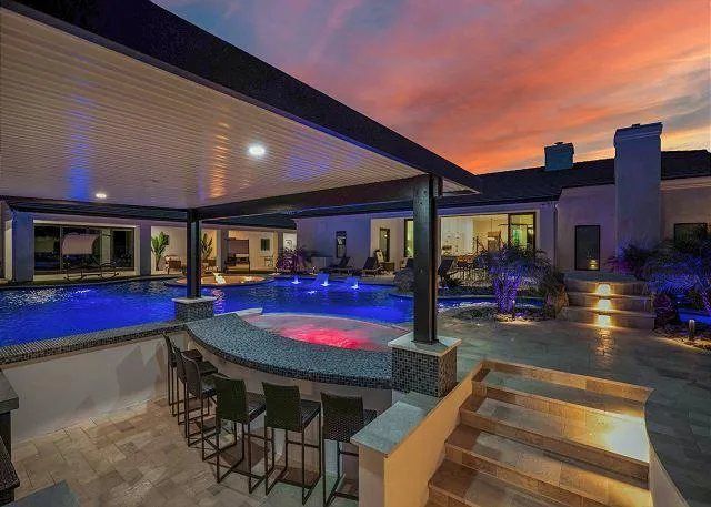 Luxury backyard with pool, bar, and outdoor seating under a pergola at sunset.