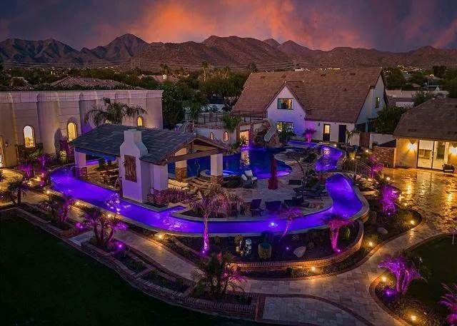 Nighttime aerial view of a luxurious property with a pool, bar, and lights in front of mountain backdrop.