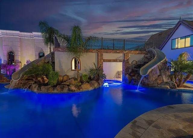 Swimming pool with slides, waterfalls, and landscaping, lit with blue lights at night.