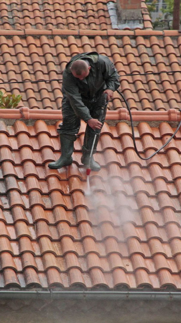Une personne en combinaison sombre nettoie à haute pression un toit de tuiles rouges.